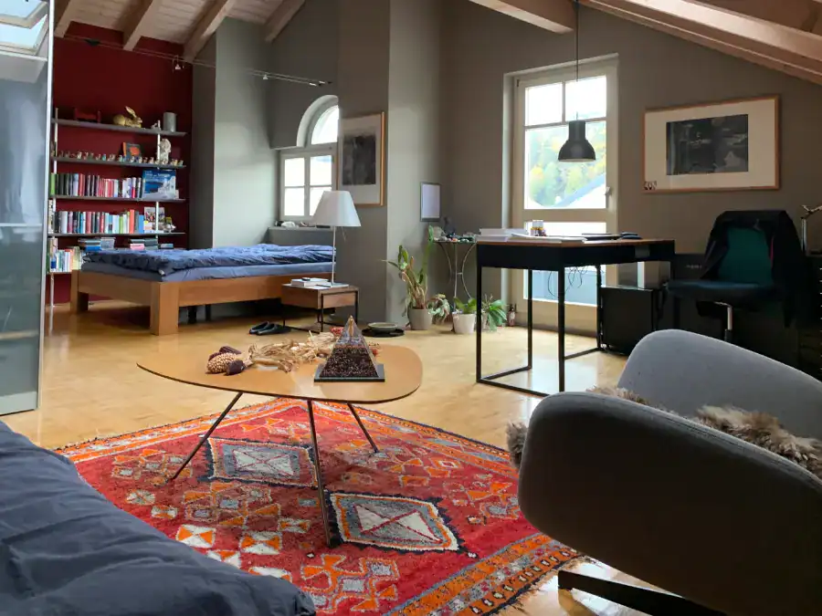 Farbgestaltung Innenraum: Ein Wohnraum im Dachgeschoss mit hoher Holzdecke in Sattelform und seitlichen Fensteröffnungen. Die Möblierung zeigt den Sitz-, Schlaf- und Arbeitsbereich. Wände in Umbra und Rouge rubis. Honigfarbener Parkett, rotoranger Orientteppich, warmfarbige Holzmöbel, ergänzt mit Details in Schwearz.,