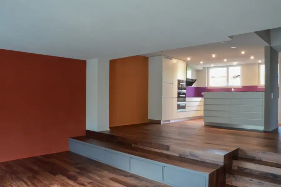 Studio für Yoga und Pilates. Ansicht des Gruppentrainingsraums vom Eingang aus in Richtung Westwand. Stirnwand in Goldocker mit links zwei offenen Türen zum Umkleideraum und zum Personal-Trainingsraum sowie rechts davon einem grossen kreisförmigen Spiegel. Angrenzende Wand rechts in Weiss mit halbhohen Fenstern. Links weisse Wand mit hellgrauer Tür zum Waschraum. Im vorderen Bereich links Stirnwand zum Waschraum in Goldocker mit einer Empfangstheke.