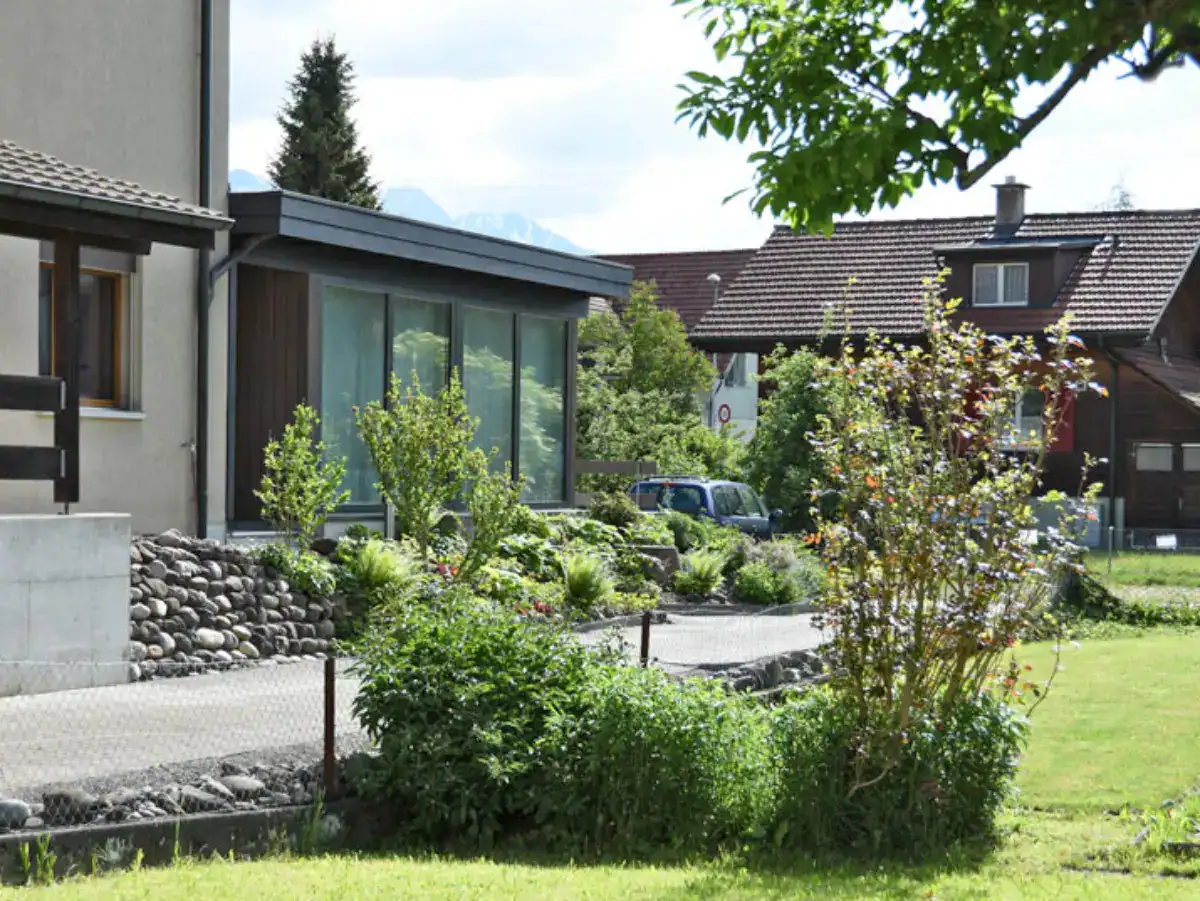 Atelier raumatmosphaeren.ch: Aussenansicht des Holzanbaus mit Eingang in frühlingshafter grüner Umgebung, erste blühende Sträucher und Tulpen, Nachbargebäude im Hintergrund mit dunkelbrauner Holzfassade.