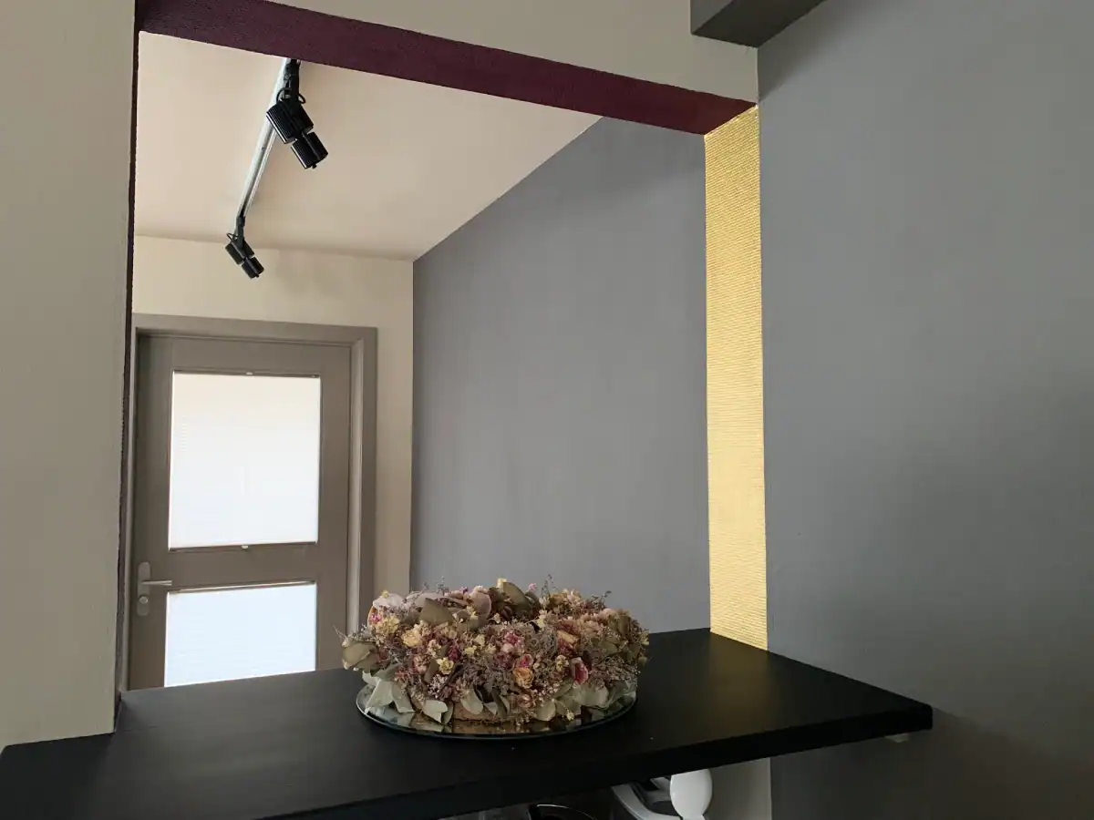 Atelier Farbgestaltung: Blick durch eine Wandöffnung mit Theke in den Eingangsbereich. Brüstung in Schwarz, Wände in warmer Sandfarbe und mittlerer Umbra, feine Details in Gold und Burgunderrot. Auf der Theke ein Kranz mit getrockneten Rosenblüten in feinem Moos und Rosatönen.