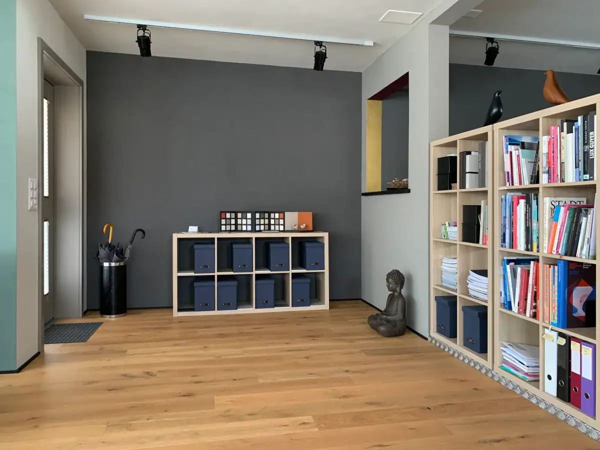Atelier Farbgestaltung: Eingangsbereich mit Büchergestell, Buddhafigur, Gestell mit Farbmustern. Warmer Eichenboden, Wände in Sandfarbe, helle und mittlere Umbra, Anthrazit, Details in Gold, Burgunderrot und Schwarz.