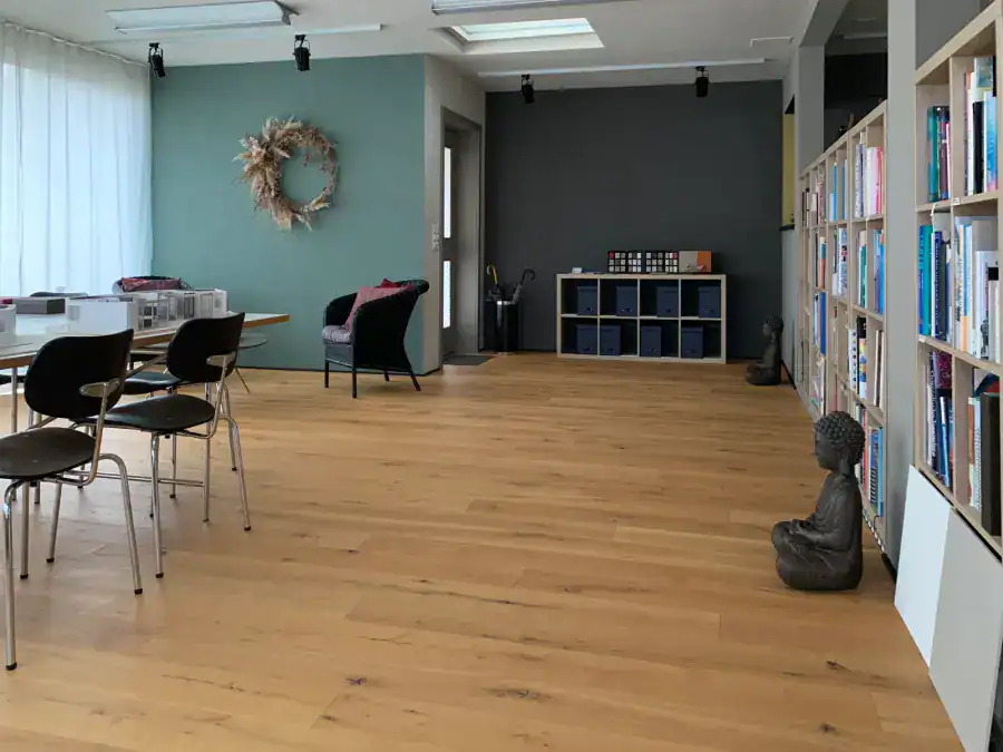 Atelier Farbgestaltung: Blick in den Innenraum beim Eingang. Die Wände sind in hellem Sandbeige, Spanischgrün und Anthrazit gehalten, rechts ein langes Büchergestell und eine Buddhafigur, links Stühle und Arbeitstisch.