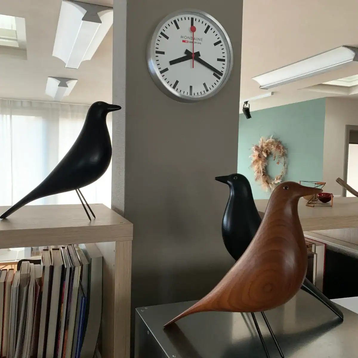 Atelier Farbgestaltung: Blick auf das hellbeige Büchergestell mit drei Design-Vögeln in Nussbaum und Schwarz, vor einer SBB-Uhr. Im Hintergrund sieht man den oberen Teil des vorderen Raums des Ateliers mit Wänden in Spanischgrün und hellem Sandbeige.