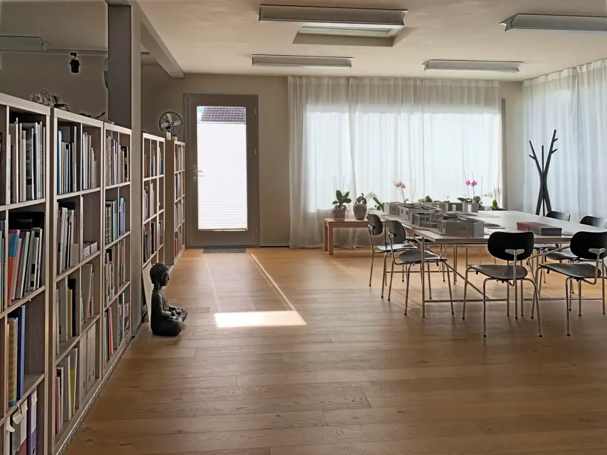 Farbgestaltung für Geschäftsräume: Blick in einen lichtvollen Raum mit einem grossen Besprechungs- und Arbeitstisch. Eichenparkett, links ein langes hellbeiges Büchergestell als Raumtrennung, grosse Fensterflächen mit hellen Vorhängen. Wandfarben: helle Umbranuance und mittlere Umbra für Türrahmen und Wandelemente.