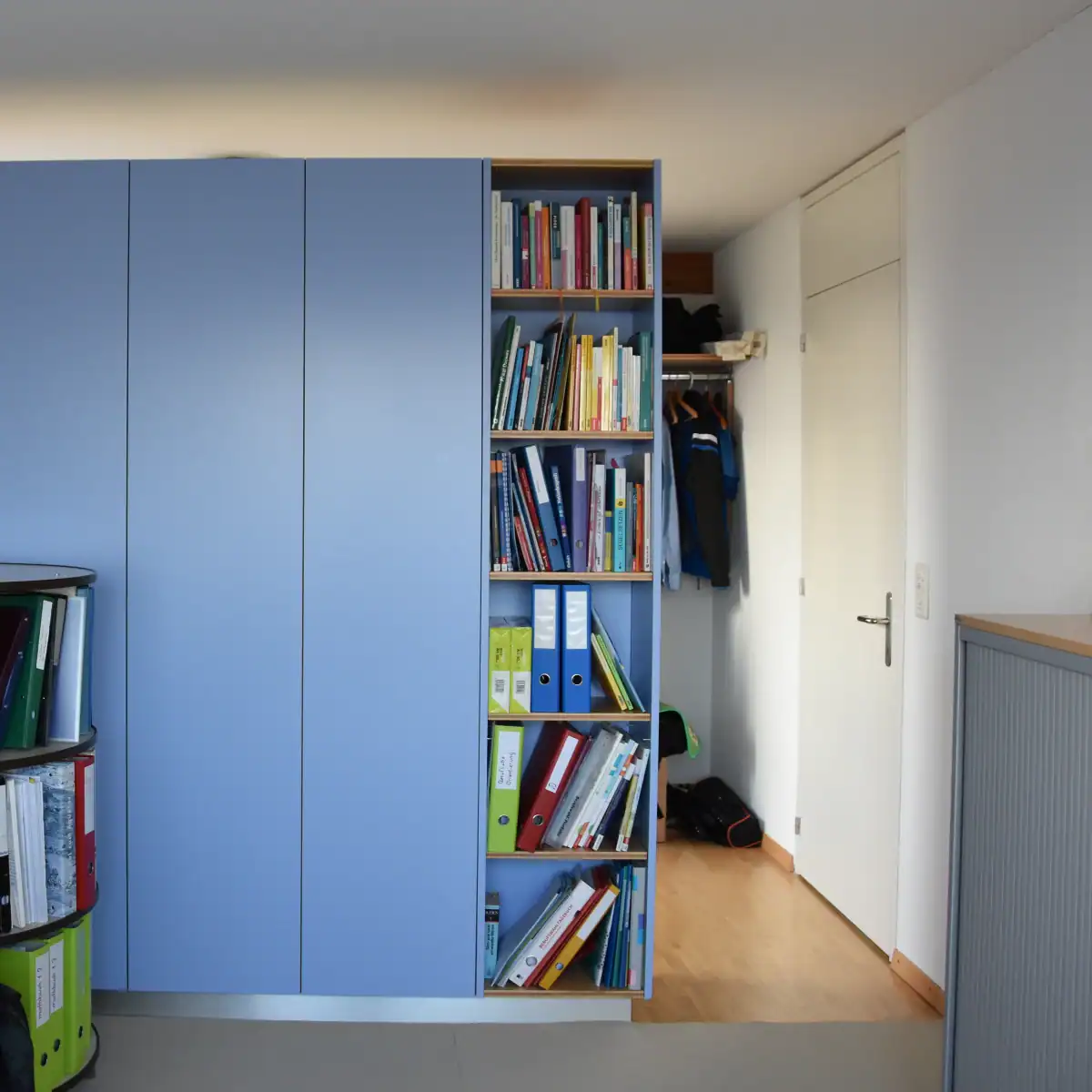 Farbgestaltung für ein Home-Office: Blick in die Ausgangslage eines Büroraums, der eine neue Wandgestaltung bekommen soll. Bestehende Farben sind die Decke, die Wände und die Tür in Weiss, das honigbraune Holzparkett, graue Büromöbel mit Holzabdeckung und ein blauer Schrank.