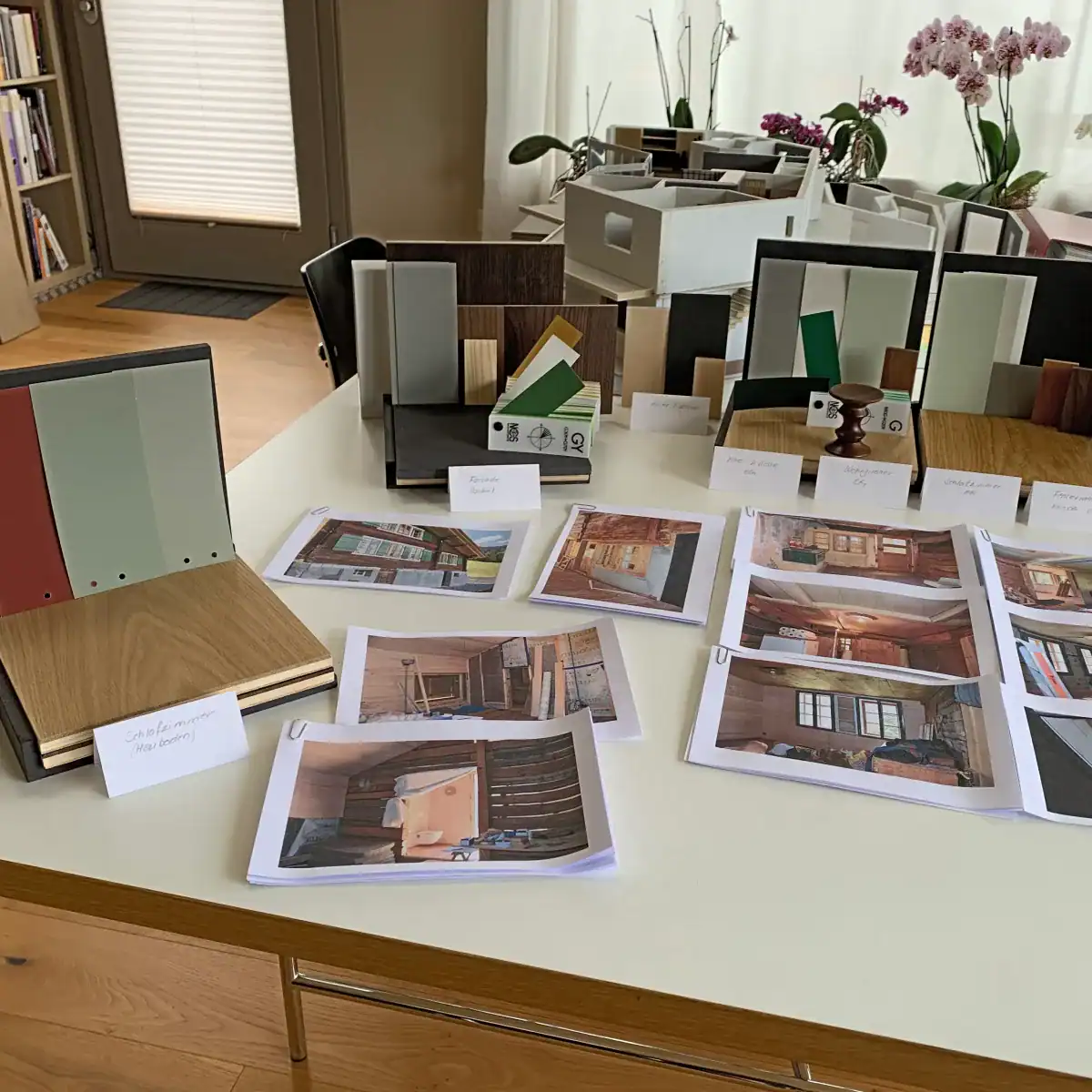 Farbgestaltung Innenraum: Das Bild zeigt einen Ausschnitt der Konzeptpräsentation auf einem Tisch mit verschiedenen kleinen Raummodellen mit Farbmustern, Farbfächern, Holz-Materialien und Fotografien. Farbpalette: Weiss, Umbra, sanfte Grünschattierungen, Akzente in Rot.