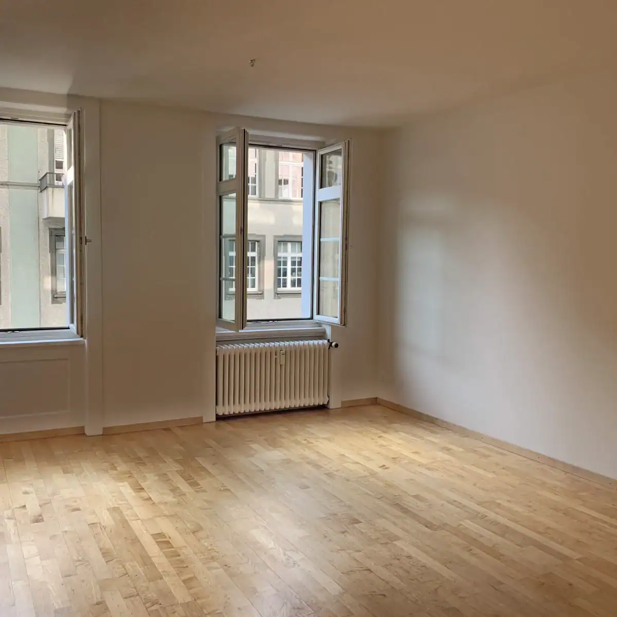 Ausgangslage für die Gestaltung eines Praxisraums in der Stadt. Die typische Situation zeigt einen kahlen Raum mit hellbraunem Laminatboden, weissen Wänden, Fenstern und Heizkörper.