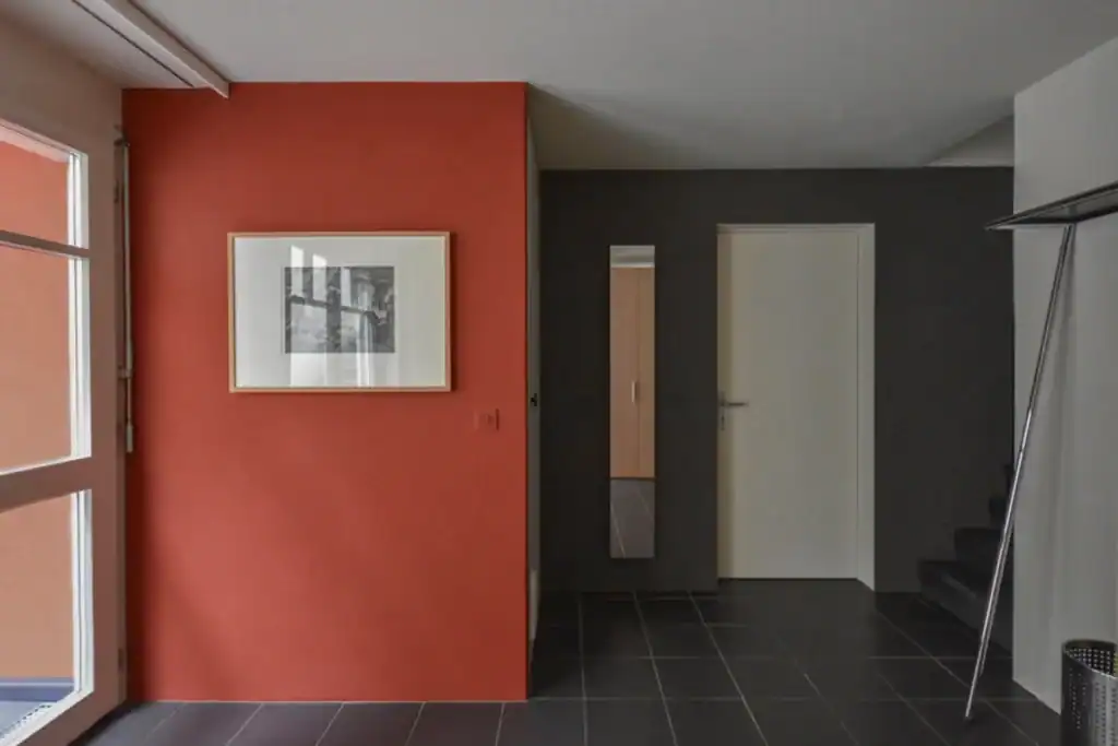 Farbgestaltung Wohnhaus: Eingangsbereich im Untergeschoss mit belebendem Farbakzent in Granatrot. Weitere Wandfarben in Anthrazit und Umbraweiss, dazu anthrazitfarbene Steingutbodenplatten.