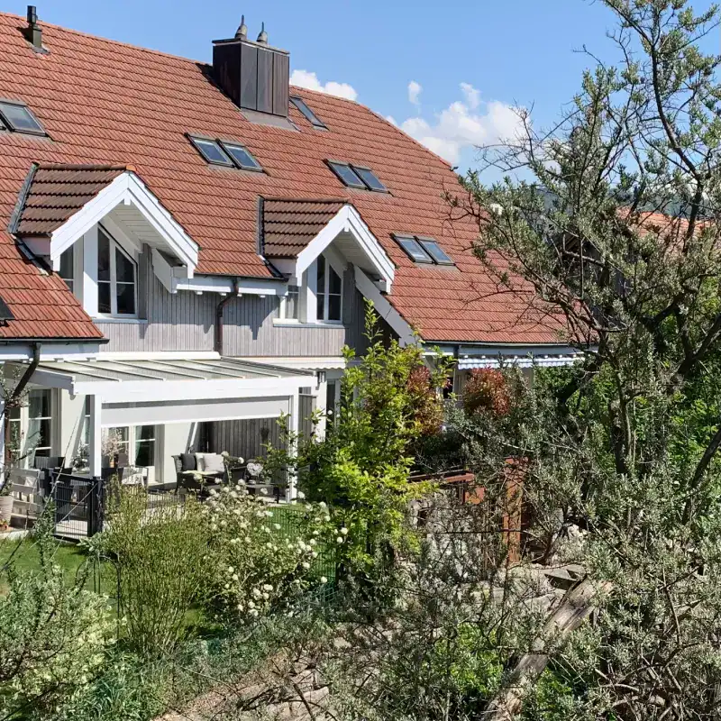 Inspirationsbild: Eine Hausfassade in Gartenumgebung. Blick auf die Fassade mit hellgrauer Holzverkleidung, ein Terrassenvordach mit Sitzplatz in weichem Weiss und weissen Fenstern, Satteldach mit rostroten Ziegeln. Blühender Gartenbereich mit Sträuchern und blauer Himmel im Hintergrund.