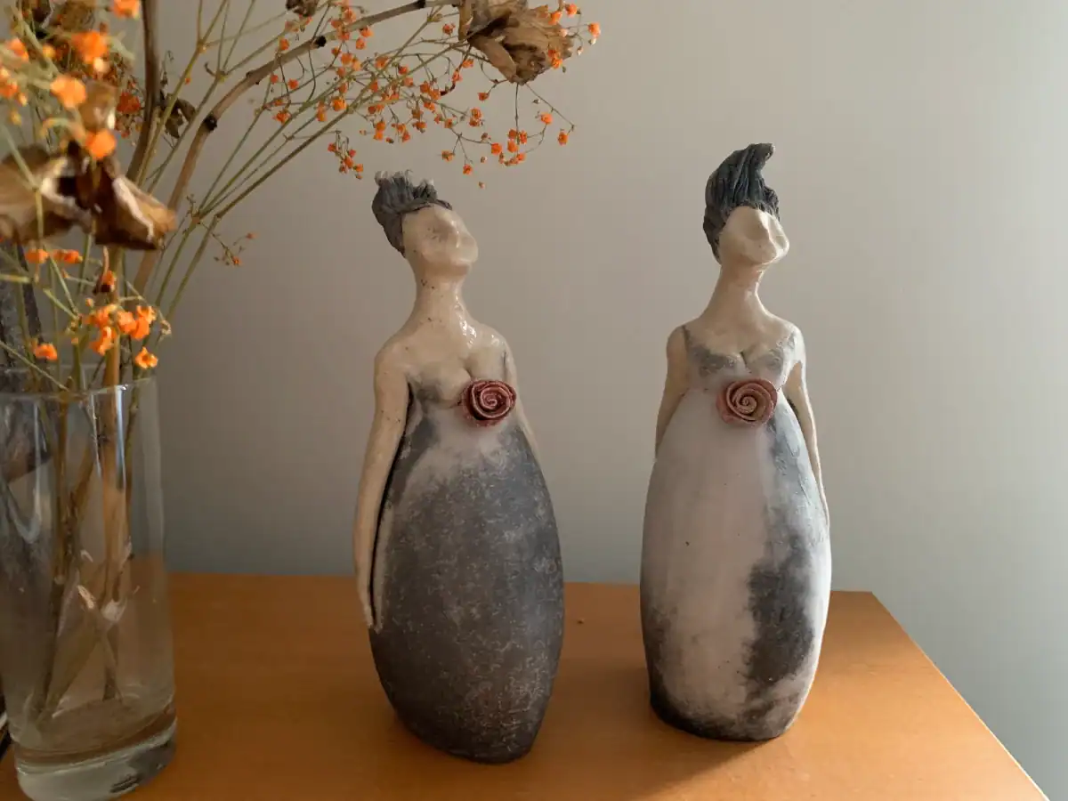 Farbgestaltung: Zwei weibliche Keramikfiguren in langem Kleid mit einer Rose als Brustornament in erdigen Farben. Seitlich getrocknete orangefarbene Blüten im Licht