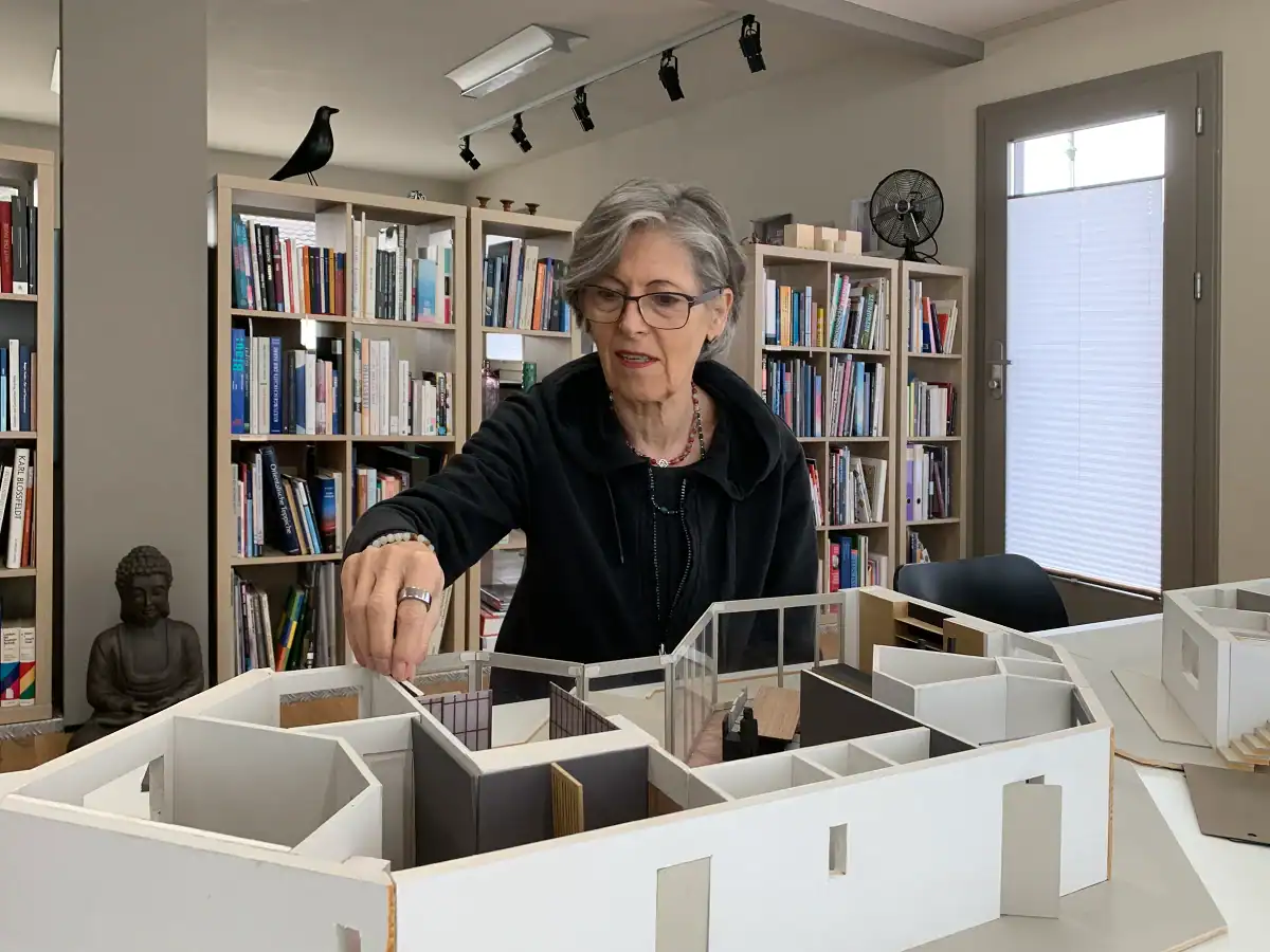 Regula Bielinski, Farbgestalterin, arbeitet an einem Modell des Erdgeschosses eines grossen Hauses. Sie testet die Wirkung eines Farbmusters im Modell. Im Hintergrund ein Büchergestell mit ihrer Farb- und Architekturbibliothek als Trennwand zum hinteren Atelierbereich.