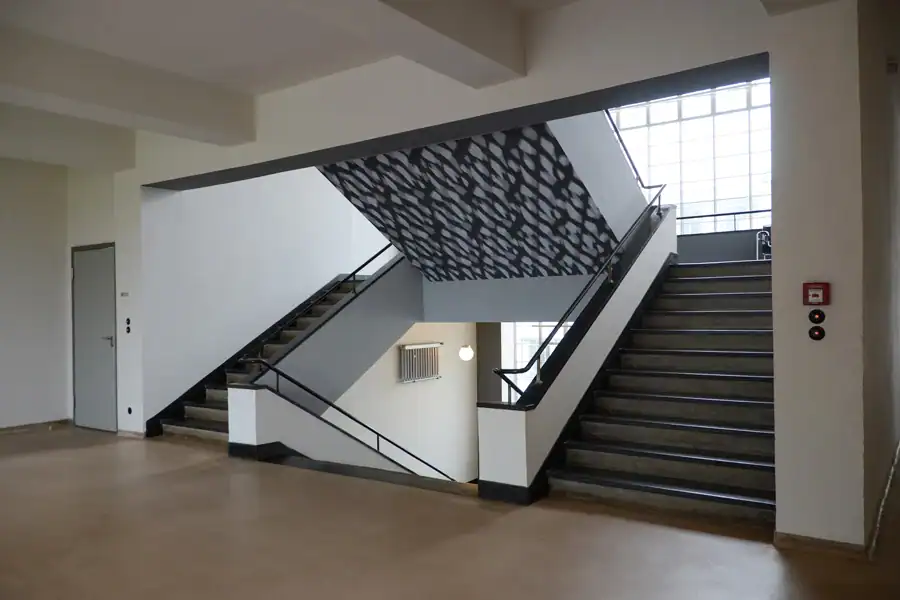 Treppenhaus eines Museums im Bauhausstil mit eingefügtem ornamentalem Element. Brauner Linoleumboden, weisse Wände, schwarze Treppenbrüstung. Auf der schrägverlaufenden Unterseite des Aufgangs ein geometrisches lineares Muster in Schwarz, Grau und Weiss mit dreidimensionaler Wirkung.