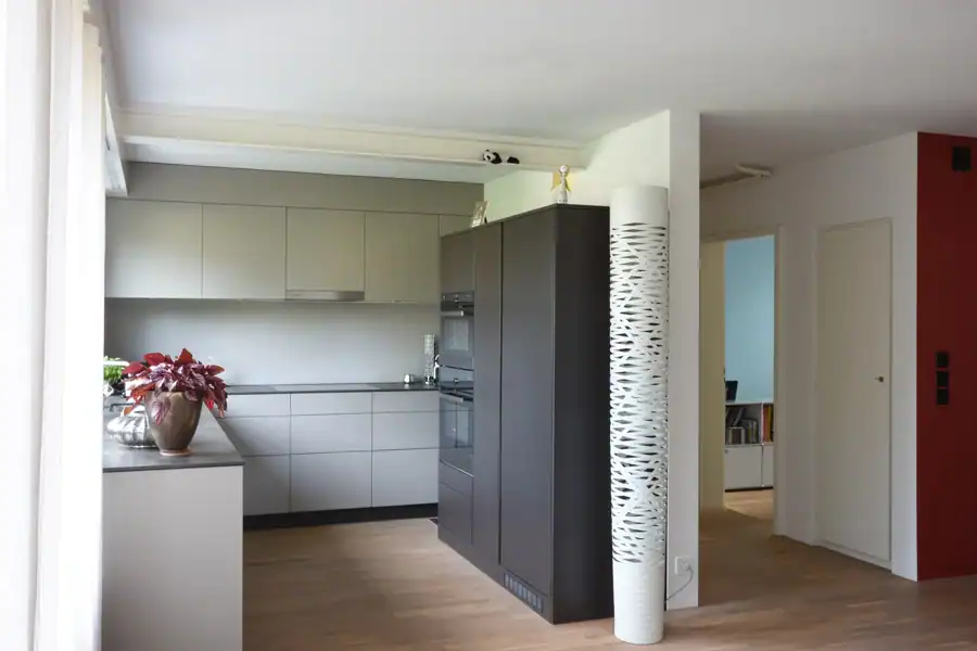 Küchenbereich und Gang mit Blick ins Schlafzimmer im Mittelgeschoss eines Einfamilienhauses aus den 50er-Jahren. Links die Küche mit hellen Fronten und anthrazitfarbenem Hochschrank, in der Mitte eine dekorative weisse Säule als Raumtrenner. Rechts der Durchgang zum Schlafzimmer mit blaugrüner Wand und die rote Akzentwand des Treppenhauses.