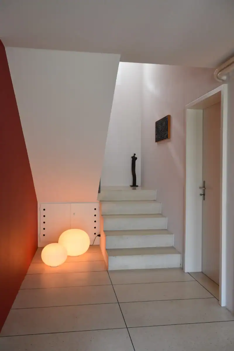 Eingangsbereich mit Treppenhaus im Einfamilienhaus aus den 50er-Jahren. Links rote Wand, hellbeige Bodenplatten und Treppentritte, übrige Wände in abgetöntem Weiss. Unterhalb der Treppe am Boden zwei Kugellampen, die eine warme Atmosphäre ausstrahlen.