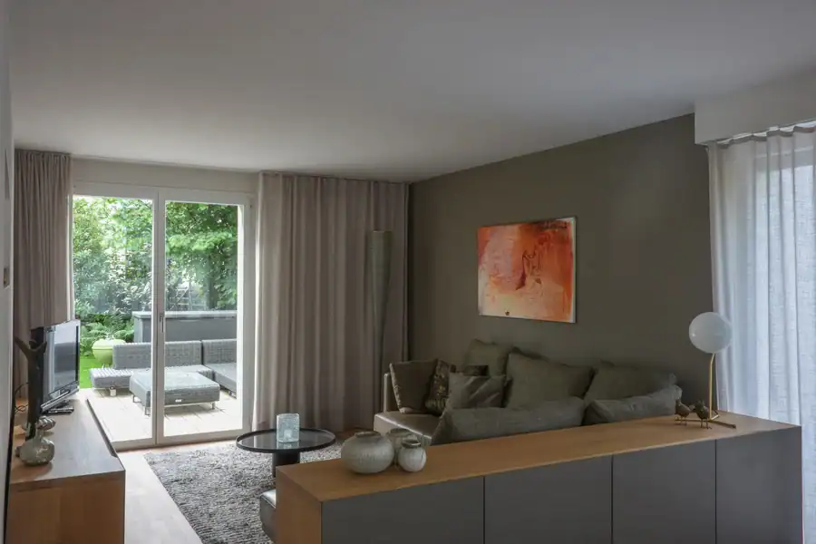 Livingbereich im Einfamilienhaus aus den 50er-Jahren. Hellbraunes Holzparkett, beige melierter Teppich. Geradeaus Blick in den Garten mit hellbeigen Vorhängen. Rückwand rechts in Terre d’Ombre moyenne (warmes Mittelbraungrau) mit einem roten abstrakten Gemälde. Beigegraues Sofa, Sideboards aus Holz mit umbrafarbenen Fronten. Verschiedene Vasen, Dekorobjekte und eine Lampe auf den Sideboards.ten.