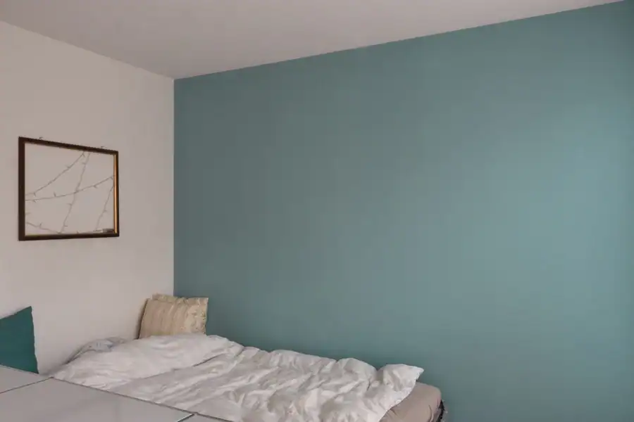 Schlafzimmer im Einfamilienhaus aus den 50er-Jahren. Raumausschnitt mit links abgetönter weisser Wand mit einem Bild, rechts gegraute blaugrüne Akzentwand. Davor Bett mit weissem Duvet und zwei beigen sowie einem blaugrünen Kissen. Ausschnitt eines weissen USM-Regalmöbels als Trennung zum vorderen Raum.