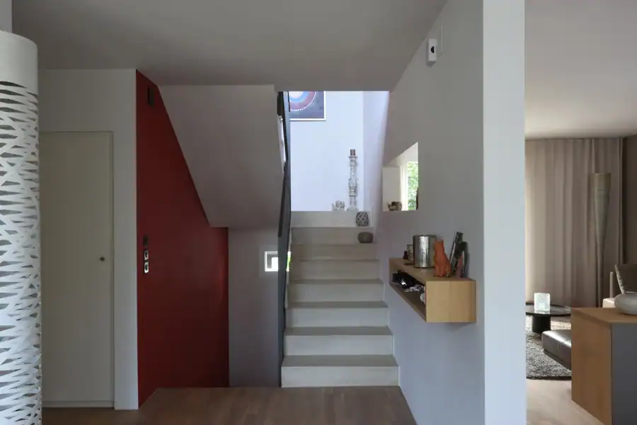 Treppenhaus im Mittelgeschoss des Einfamilienhauses aus den 50er-Jahren. Links Akzentwand in Rot, die vom Eingang bis ins Obergeschoss leitet, übrige Wände in abgetöntem Weiss. Treppentritte in hellbeigen Steinplatten, schwarze Metallbrüstung. An der rechten Wand ein längliches Holzregal auf mittlerer Höhe. Weiter oben ein schmaler, waagrechter Wanddurchbruch mit Sicht ins Wohnzimmer. Rechts schmaler Blick in den Livingbereich. Verschiedene Skulpturen und Dekorelemente.