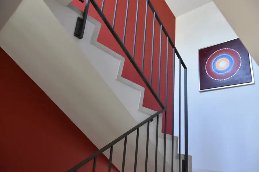 Detail der Treppenhausgestaltung im Einfamilienhaus aus den 50er-Jahren. Schräge Sicht nach oben ins Obergeschoss. Links Akzentwand in Rot, die vom Eingang bis ins Obergeschoss leitet, übrige Wände in abgetöntem Weiss. Treppentritte in hellbeigen Steinplatten, Betonunterteil in abgetöntem Weiss, schwarze Metallbrüstung. An der Rückwand ein quadratisches Bild mit kreisförmigem Motiv in Weiss, Orange und Blau auf tiefdunklem rotviolettem Hintergrund. Klare, reduzierte Architektursprache, die aus den Kontrasten lebt.