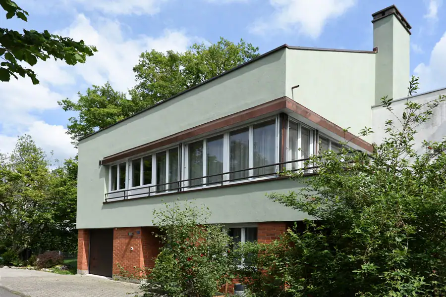Salbeigrün-graue Fassade eines Einfamilienhauses aus den 50er-Jahren in grüner Quartierumgebung mit Backsteinsockelgeschoss und langgezogener Fensterfront mit Kupferabschlüssen und -brüstung.
