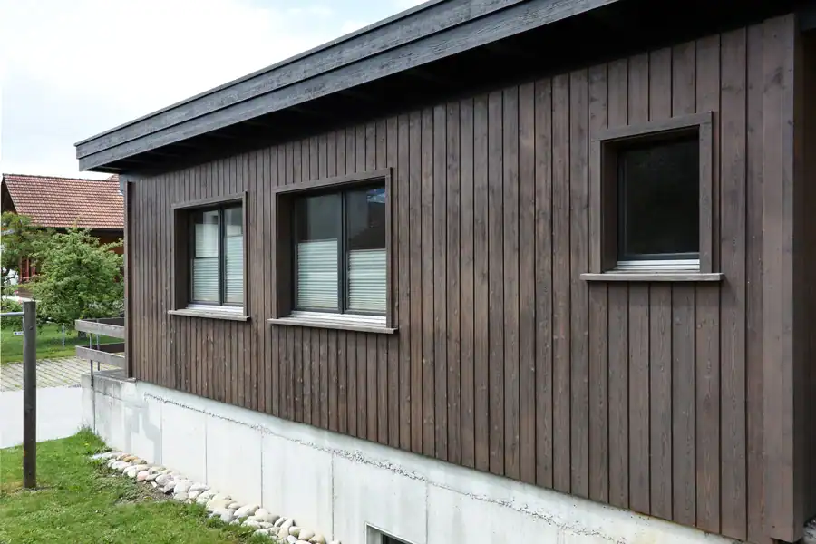 Renovierte Holzfassade eines eingeschossigen Ateliers für Farbgestaltung. Südseite. Hellgrauer Betonsockel, dunkelbraune Holzverschalung, Flachdach mit anthrazitfarbenem Vordach.