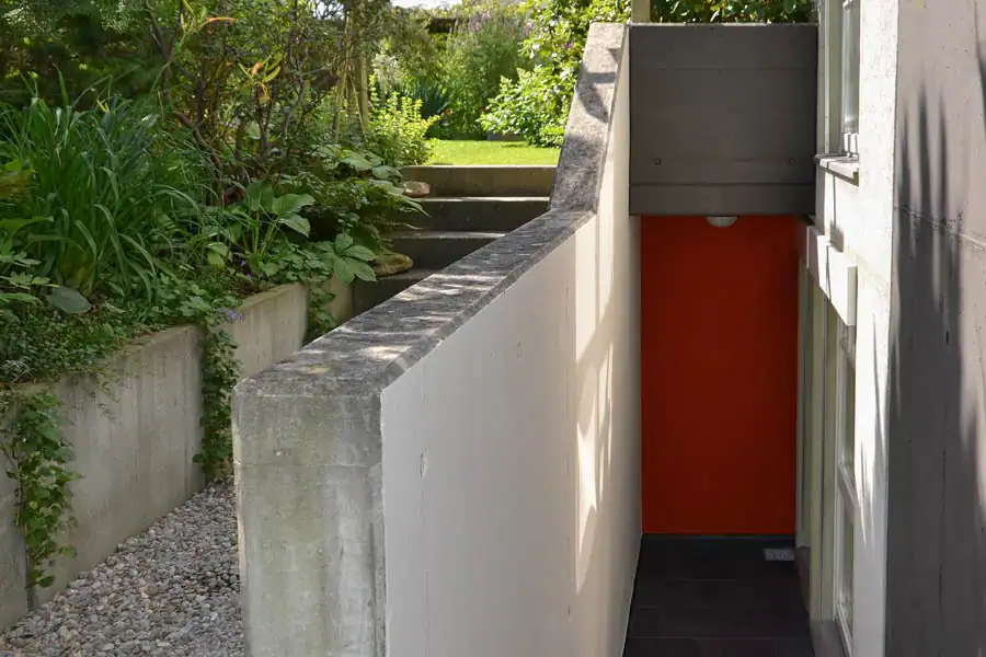 Äusserer Eingangsbereich eines Wohnhauses. Schmale Treppe ins UG neben dem Gartenzugang, links Betonwand, rechts Hausfassade mit Fensterfronten. Grünlichweisse Fassade, weisse Fensterrahmen, Betonflächen in heller und dunkler Umbra, Rückwand neben Tür als granatroter Akzent.