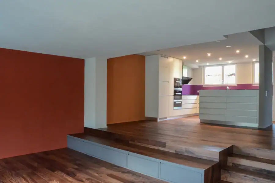 Renovation eines Wohnhauses. Offener Innenraum mit Wohn-, Essbereich und Küche. Dunkelbrauner Parkettboden, Küchenkombination in Weiss und Silbergrau, Wände in gebranntem Ocker und Goldockerorange, waagrechter fuchsiavioletter Streifen über der Küchenarbeitsfläche. Unmöblierter Zustand.