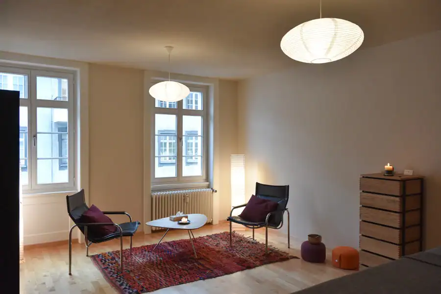 Praxisraum für spirituelles Coaching und Heilung. Warme Atmosphäre des Besprechungsbereichs mit zwei bequemen Stühlen und kleinem Tisch, Holz-Sideboard. Honigwarmes Parkett mit einem Teppich in warm rötlichen Farben. Angenehme Beleuchtung mit Japan-Papierleuchten.
