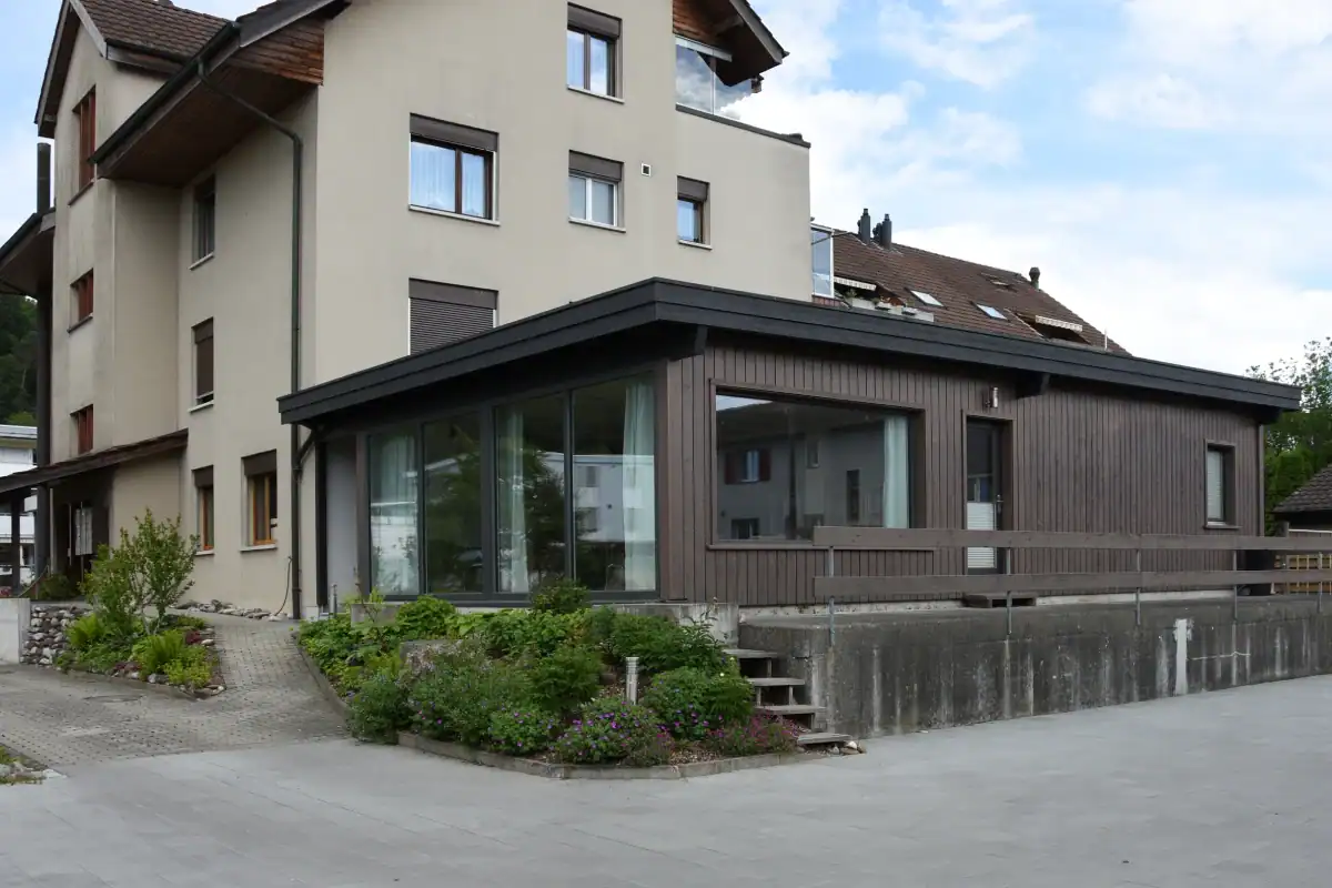 Atelieranbau mit Holzfassade: Blick auf die Nordwestseite mit Eingang und grosser Fensterfront und Westfront mit erhöhter Terrasse.