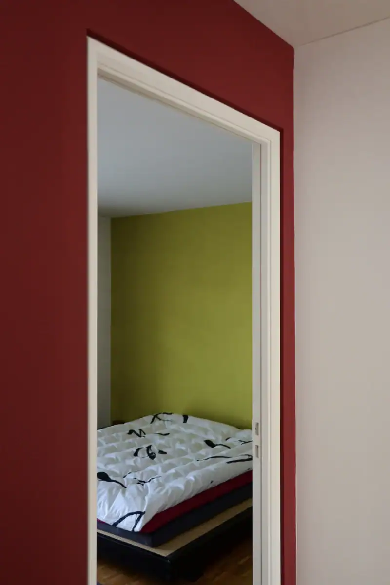 Schlafzimmer Gestaltung. Blick zur Nordostecke mit der Nische. Rechts ist ein Teil des hochwertigen dichten Vorhanges mit Silber-Bronze-Ornamenten. Links die helle Muschelkalkwand mit einer grossen hochformatigen Nische in Herbstgold, deren Boden im selben dunkelnbrauen Holz wie der Parkettboden beschaffen ist.