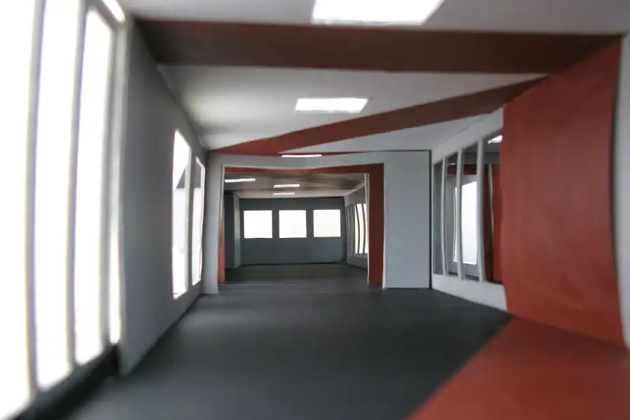 Treppenhaus im Mittelgeschoss des Einfamilienhauses aus den 50er-Jahren. Links Akzentwand in Rot, die vom Eingang bis ins Obergeschoss leitet, übrige Wände in abgetöntem Weiss. Treppentritte in hellbeigen Steinplatten, schwarze Metallbrüstung. An der rechten Wand ein längliches Holzregal auf mittlerer Höhe. Weiter oben ein schmaler, waagrechter Wanddurchbruch mit Sicht ins Wohnzimmer. Rechts schmaler Blick in den Livingbereich. Verschiedene Skulpturen und Dekorelemente.