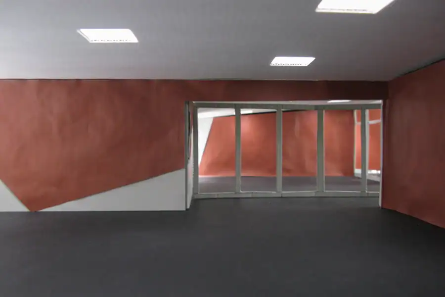 Treppenhaus im Mittelgeschoss des Einfamilienhauses aus den 50er-Jahren. Links Akzentwand in Rot, die vom Eingang bis ins Obergeschoss leitet, übrige Wände in abgetöntem Weiss. Treppentritte in hellbeigen Steinplatten, schwarze Metallbrüstung. An der rechten Wand ein längliches Holzregal auf mittlerer Höhe. Weiter oben ein schmaler, waagrechter Wanddurchbruch mit Sicht ins Wohnzimmer. Rechts schmaler Blick in den Livingbereich. Verschiedene Skulpturen und Dekorelemente.