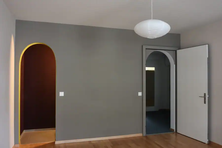 Studio für Yoga und Pilates. Personal-Trainingsraum. Blick von der Nordwand Richtung Akzentwand in Mauveanthrazit. Linke Wand zum Gruppentrainingsraum in Weiss, Tür hellgrau. Rechts Westwand in Weiss mit grossen Fenstern. Mitteldunkler honigfarbener Laminatboden und helle Holzdecke. Im Raum stehen verschiedene Pilates-Trainingsgeräte in hellem Holz und mit mittelgrauen gepolsterten Liege- und Sitzflächen. Die Anthrazitwand gibt dem Raum Halt und klare Orientierung.