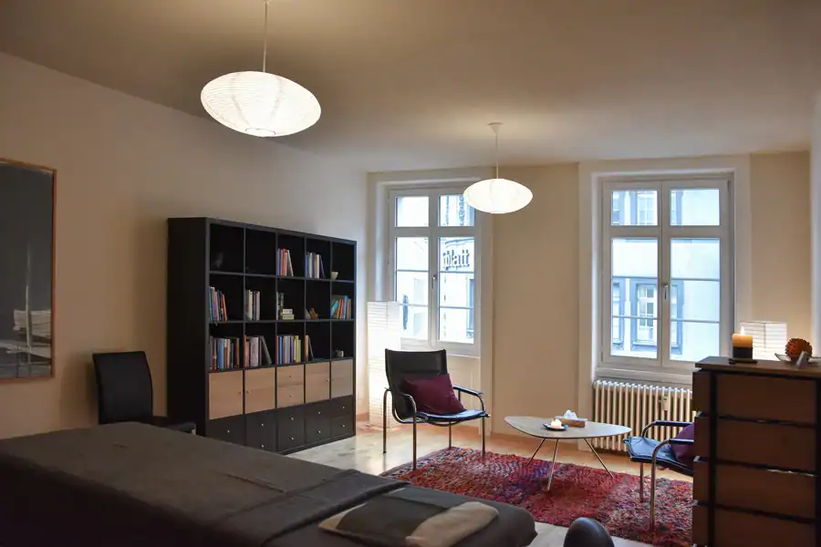 Studio für Yoga und Pilates. Ansicht des Gruppentrainingsraums vom Eingang aus in Richtung Westwand. Stirnwand in Goldocker mit links zwei offenen Türen zum Umkleideraum und zum Personal-Trainingsraum sowie rechts davon einem grossen kreisförmigen Spiegel. Angrenzende Wand rechts in Weiss mit halbhohen Fenstern. Links weisse Wand mit hellgrauer Tür zum Waschraum. Im vorderen Bereich links Stirnwand zum Waschraum in Goldocker mit einer Empfangstheke.