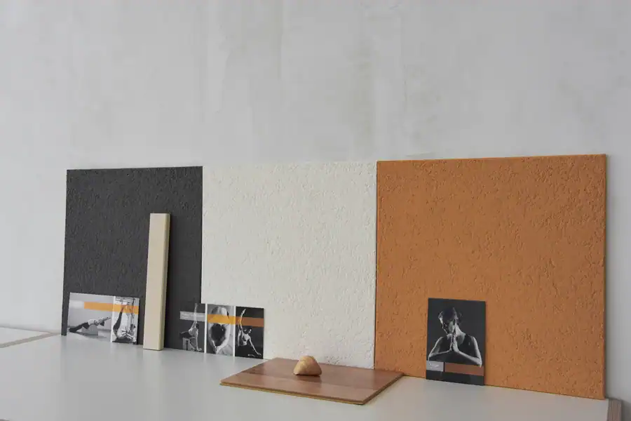 Studio für Yoga und Pilates. Bemusterung des Farbkonzepts für die neue Wandgestaltung mit grossen Musterplatten im Zusammenspiel mit den bestehenden Farben (weisse Fensterrahmen, Laminatboden und Werbepostkarten des Studios). Die grossen Musterplatten (40 × 40 cm) zeigen den entwickelten Farbklang Wollweiss, Goldocker und Mauveanthrazit. Die Platten sind auf einem tiefen weissen Regal an einer neutralen Wandfläche in gräulichem Weiss präsentiert.
