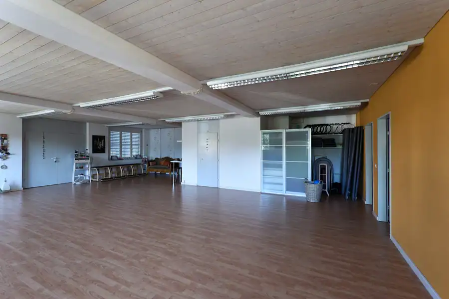 Studio für Yoga und Pilates. Mauveanthrazitfarbene Wand im Umkleideraum. Davor ein helles Holzregalmöbel mit Postkarten und Prospekten. Rechts eine hochformatige weisse Reispapier-Stehleuchte. Über dem Regal ein goldgerahmter Spiegel.