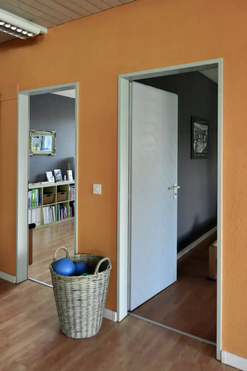 Studio für Yoga und Pilates. Blick vom Gruppentrainingsraum durch die Türen in den Umkleideraum und in den Personal-Trainingsraum. Die Wand mit den Türen ist in strahlendem Goldocker, die sichtbaren Akzentwände in den beiden Nebenräumen in Mauveanthrazit. Türrahmen und Türblätter in Hellgrau. Wirkungsvolle Differenzierung zwischen Hellgrau, Goldocker und dunklem Mauveanthrazit.